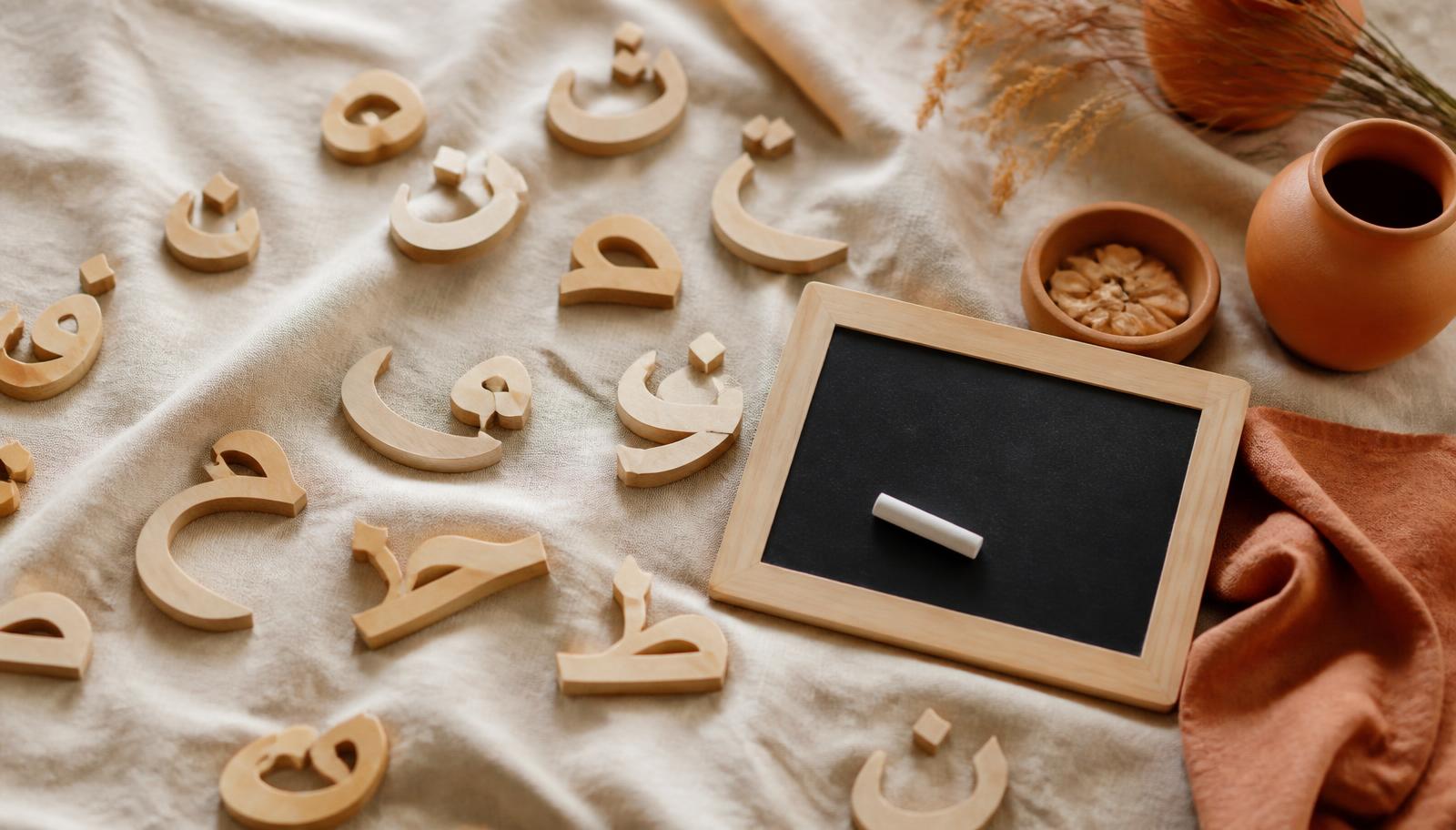 Lettres de l'alphabet arabe en bois clair posées sur du lin crème, avec une petite ardoise et une craie